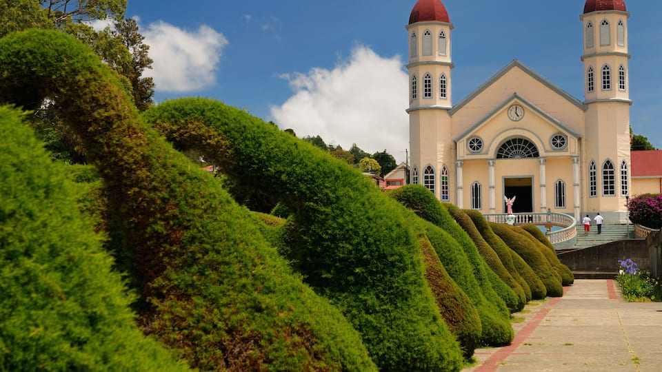 A church with two domes, a red roof, and a pathway lined with neatly trimmed bushes.
