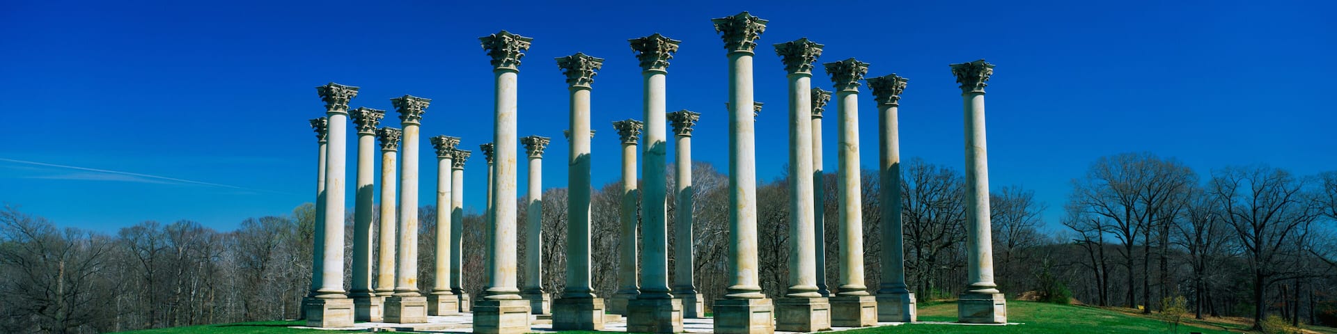 These are the historic National Capitol Columns at the National Arboretum. There is a stone walkway leading up to them surrounded by lawn against a blue sky.