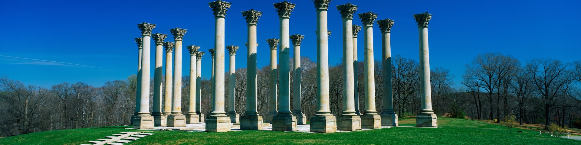 These are the historic National Capitol Columns at the National Arboretum. There is a stone walkway leading up to them surrounded by lawn against a blue sky.