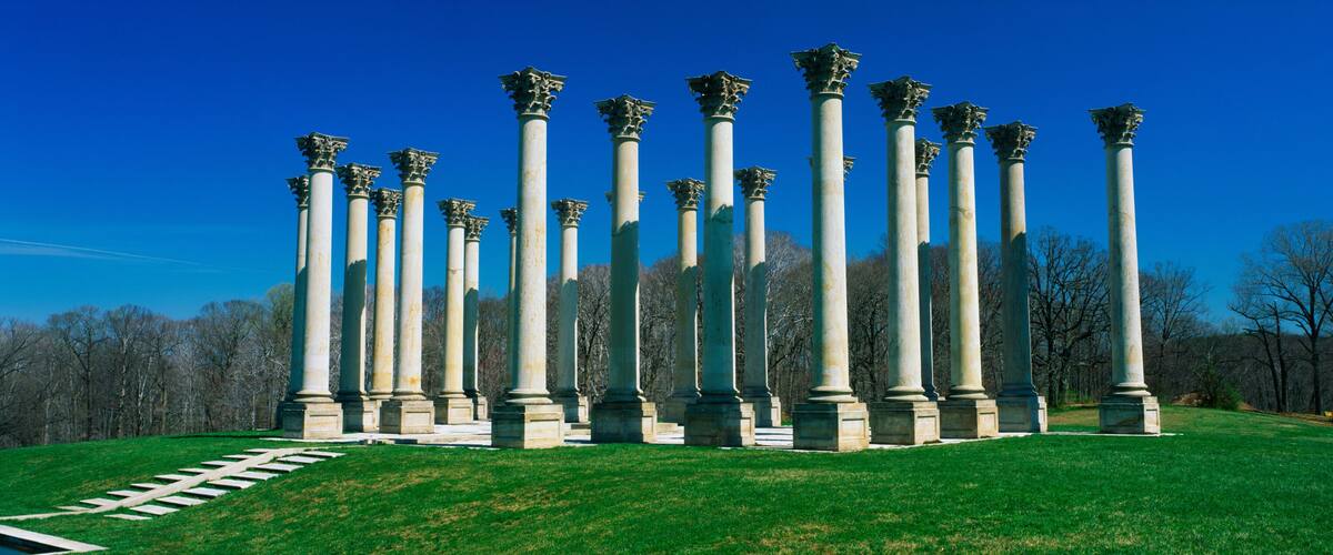 A series of tall columns with ornate capitals, arranged in a circular pattern on a grassy hill, with a clear blue sky above.