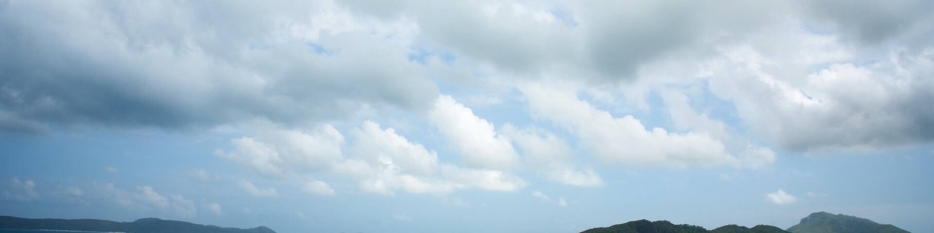 Paisagem costeira com águas azul-turquesa, uma praia de areia e montanhas distantes sob um céu nublado.