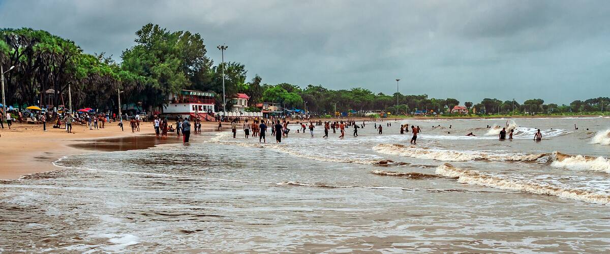 A crowded beach with waves crashing onto the shore, palm trees, and a building with a red roof.