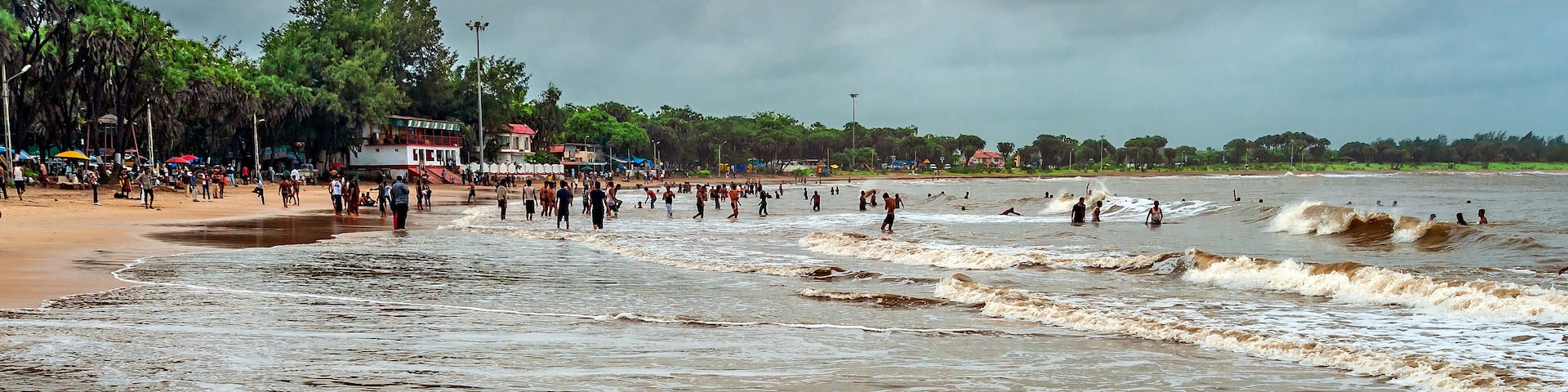 A crowded beach with waves crashing onto the shore, palm trees, and a building with a red roof.