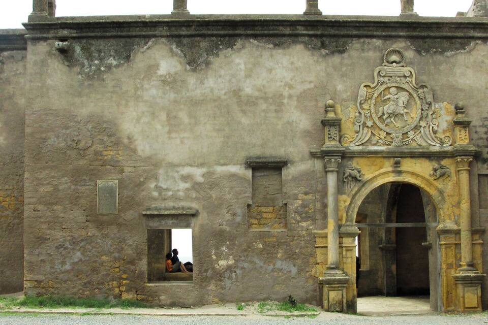 An old stone building with a weathered facade, a decorative archway, and a small window with a person inside.