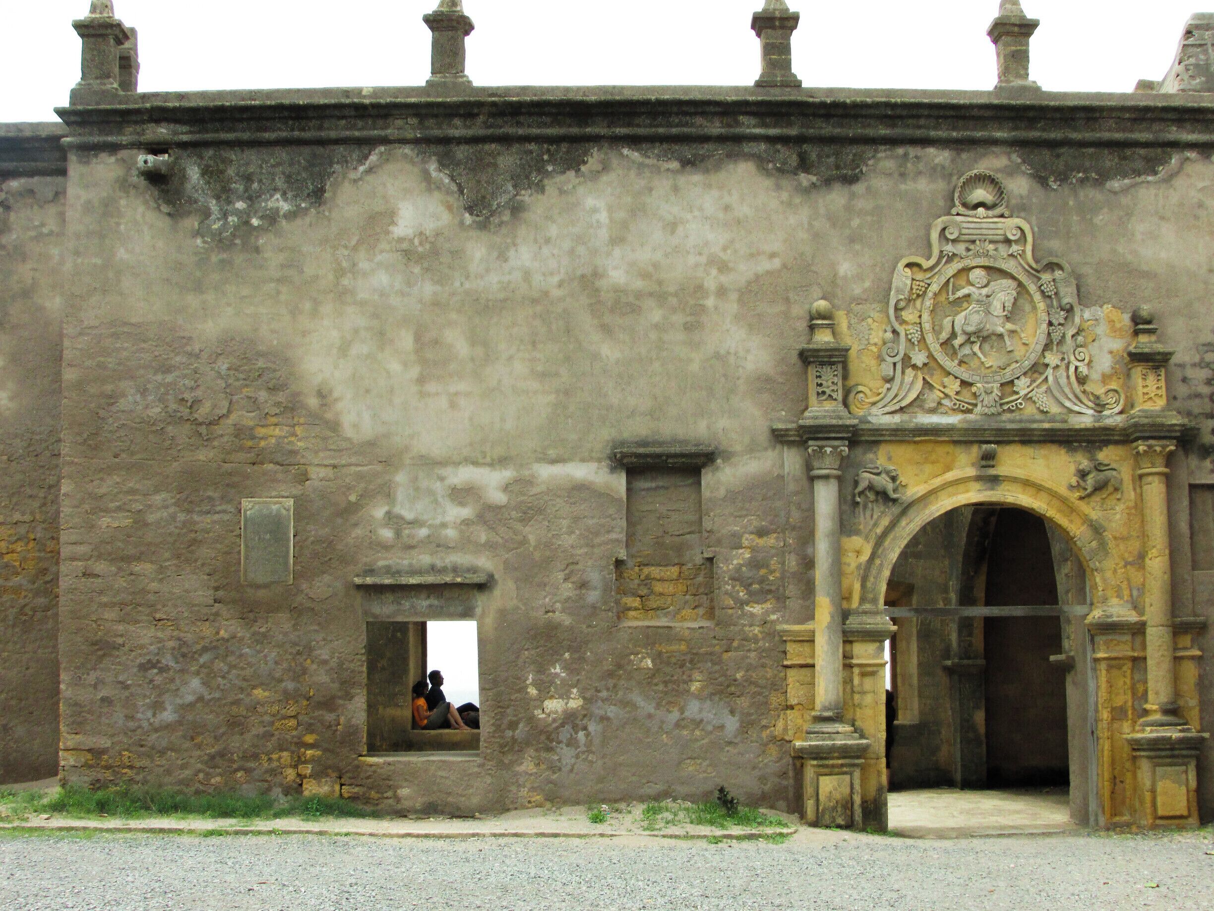 An old stone building with a weathered facade, a decorative archway, and a small window with a person inside.