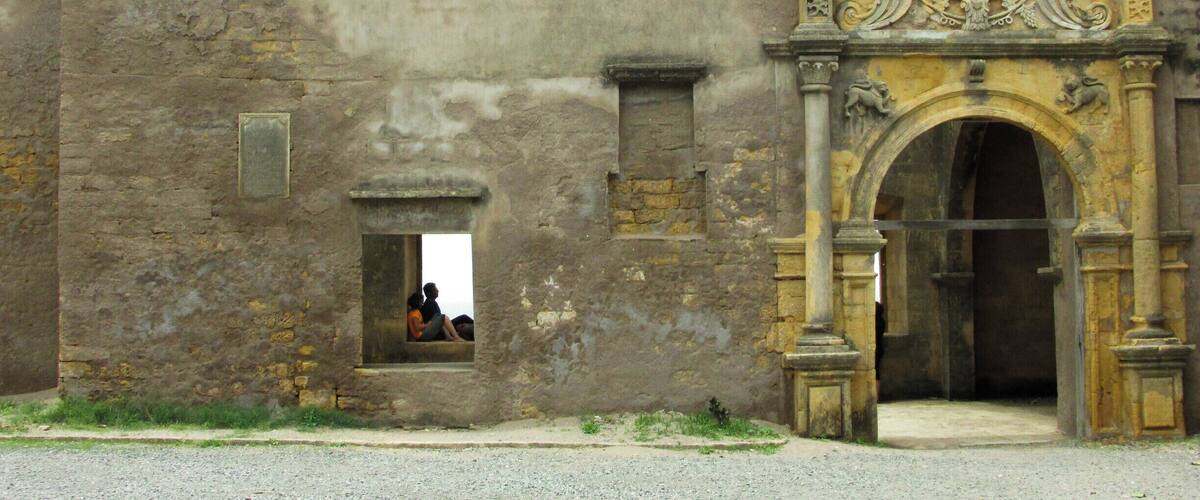 An old stone building with a weathered facade, a decorative archway, and a small window with a person inside.