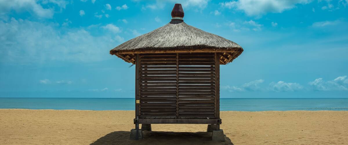 A beach hut with a thatched roof on a sandy beach with a clear blue sky.