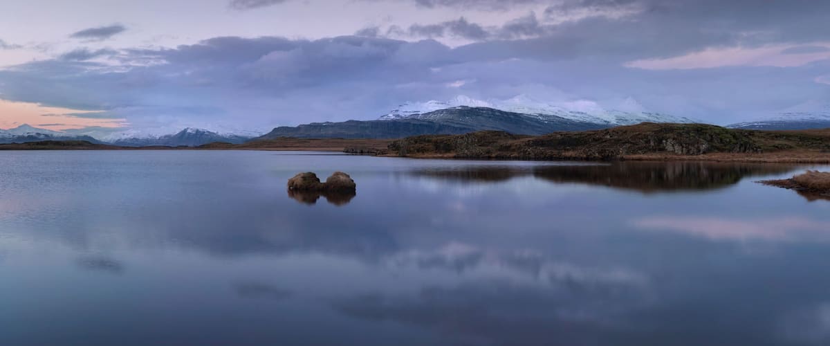 A calm lake with a reflection of a rocky outcrop and snow-capped mountains in the background.