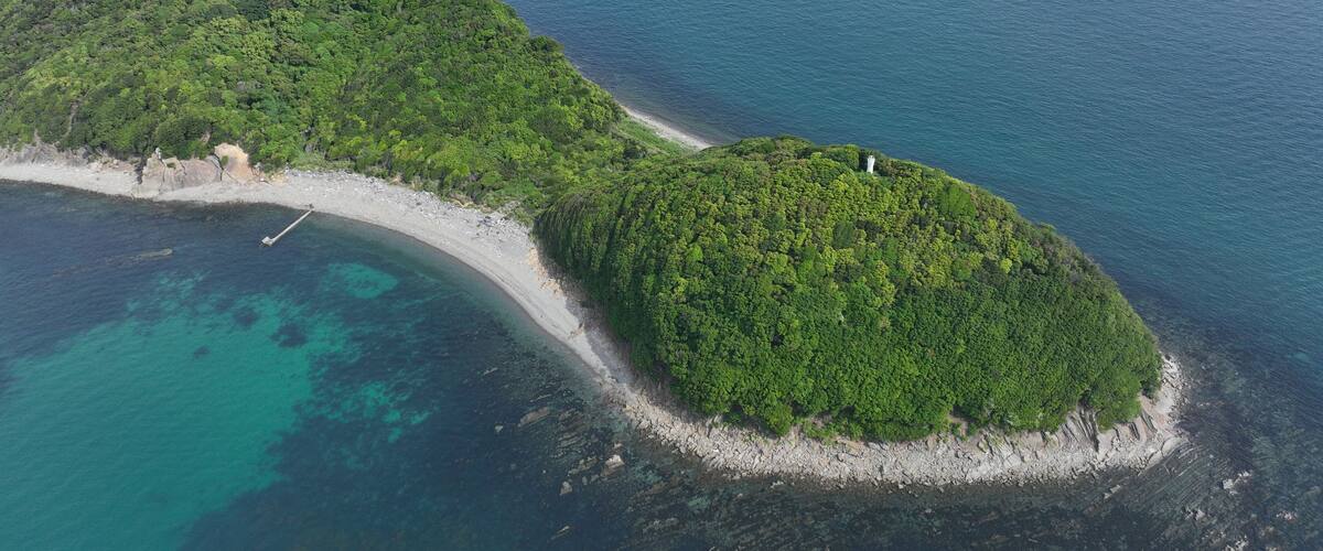 An aerial view of a lush, green island with a narrow beach and a lighthouse.