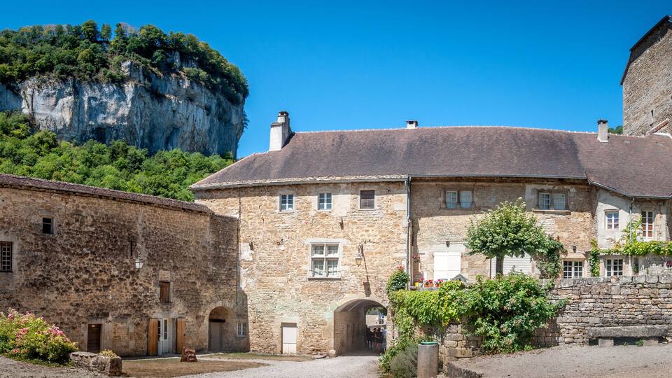 A historic stone building with a steep roof, surrounded by lush greenery and a prominent cliff.