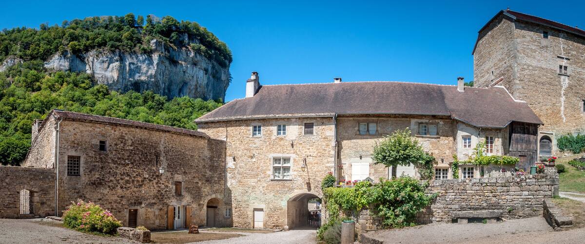 A historic stone building with a steep roof, surrounded by lush greenery and a prominent cliff.