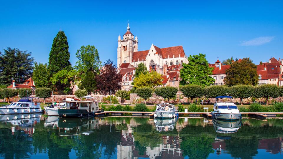 A historic building with a tower, a calm waterway with boats, and lush greenery.