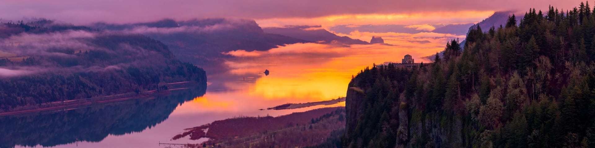 A mountainous landscape with a lake reflecting the sunset, a bridge, and a building on a cliff.