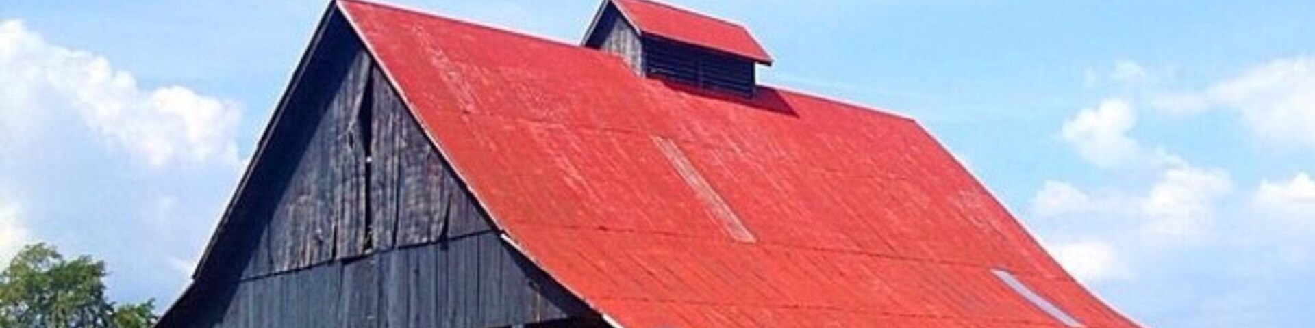 A large barn with a red roof and a dark wooden structure in a grassy field.
