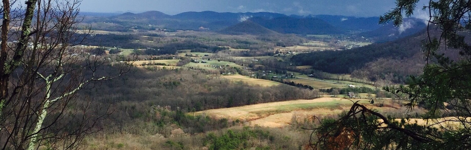 Paisagem montanhosa com um vale, áreas arborizadas e uma vista distante de uma cidade.