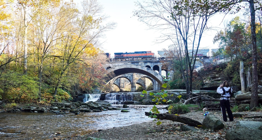 A river with a waterfall, a stone bridge, and autumn foliage.