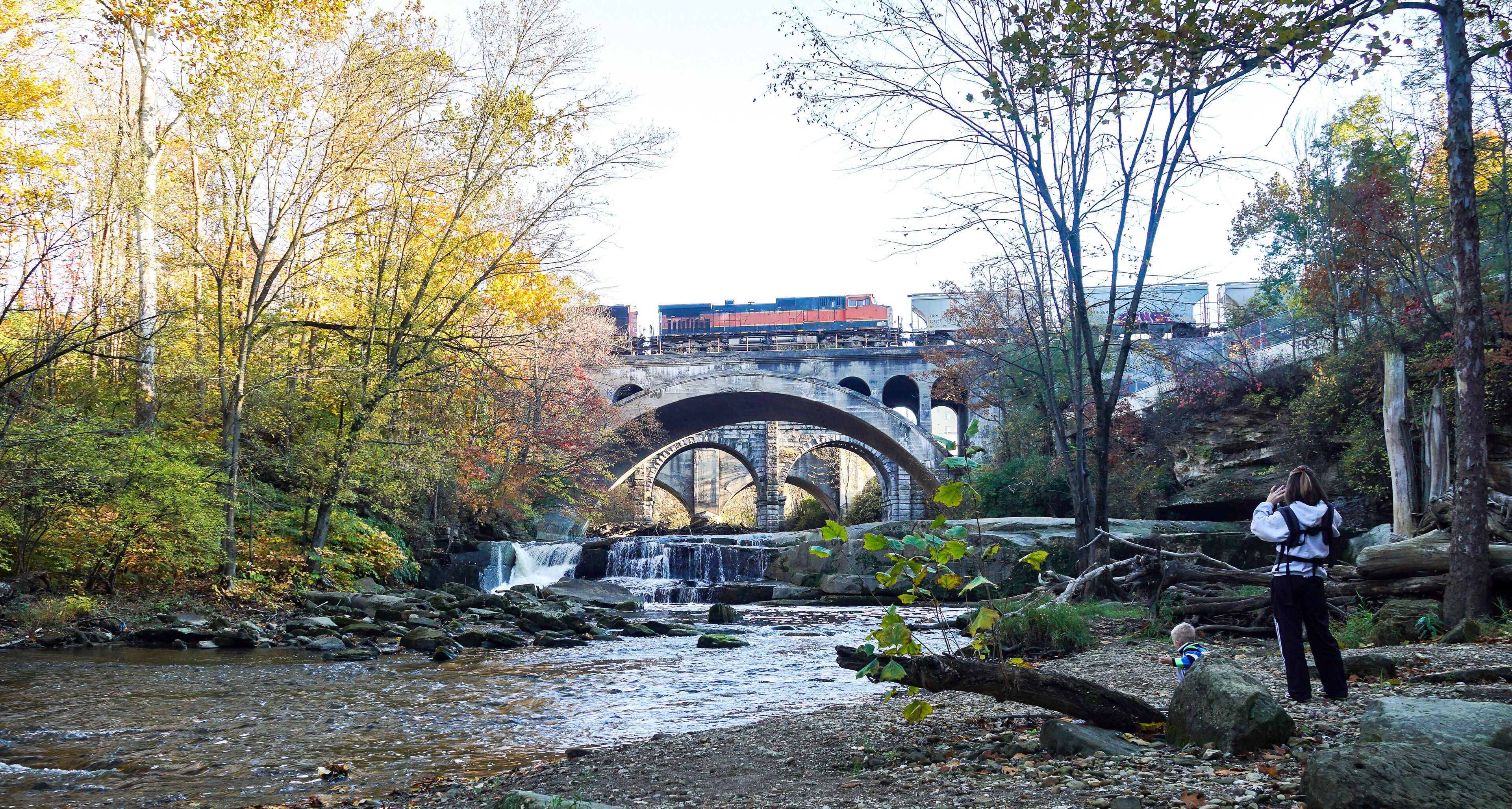 A river with a waterfall, a stone bridge, and autumn foliage.