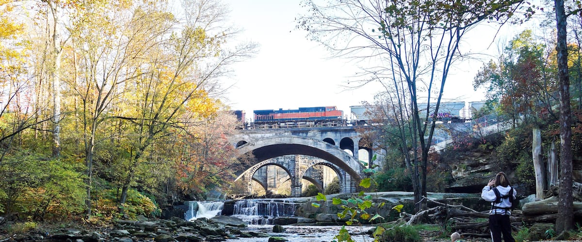 A river with a waterfall, a stone bridge, and autumn foliage.