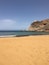 A sandy beach with clear blue water, rocky cliffs, and a small building on top.