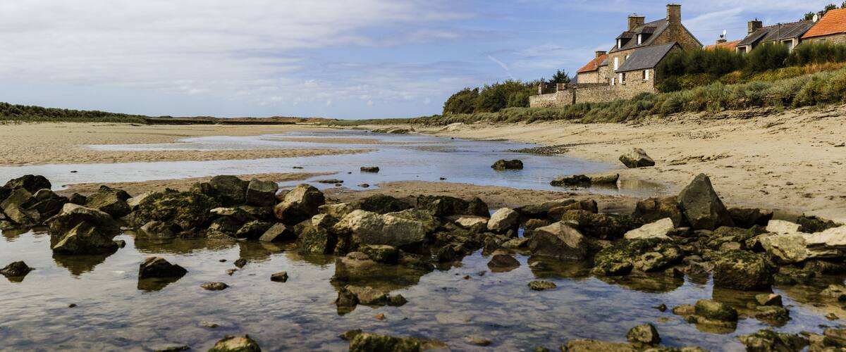 A rocky shoreline with clear water, a sandy beach, and a stone house with a red roof.