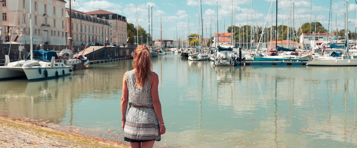 A woman standing by a marina with boats and buildings in the background.
