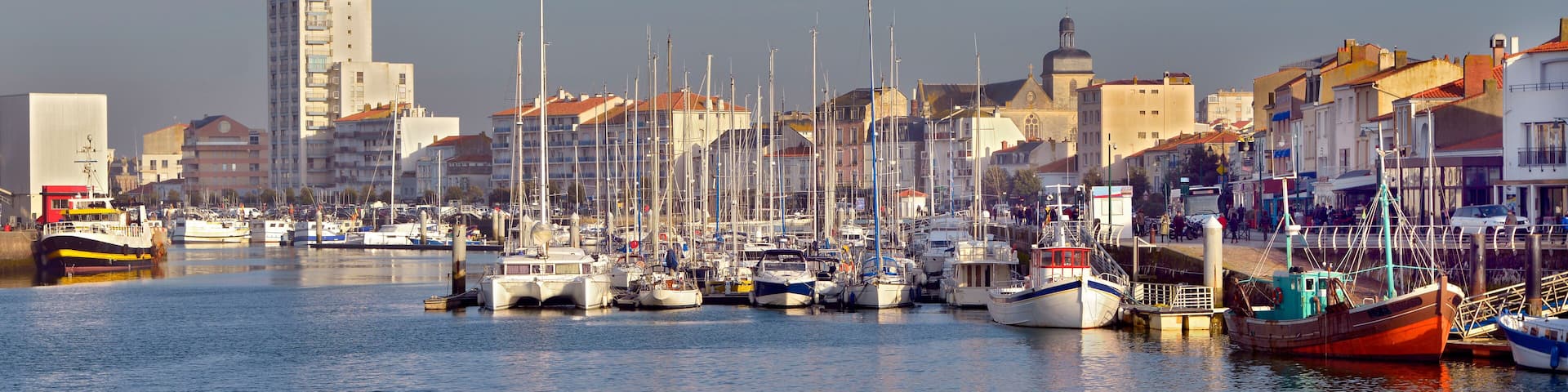 A marina with various boats docked, surrounded by buildings and a clear sky.
