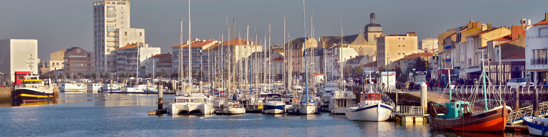 A marina with various boats docked, surrounded by buildings and a clear sky.