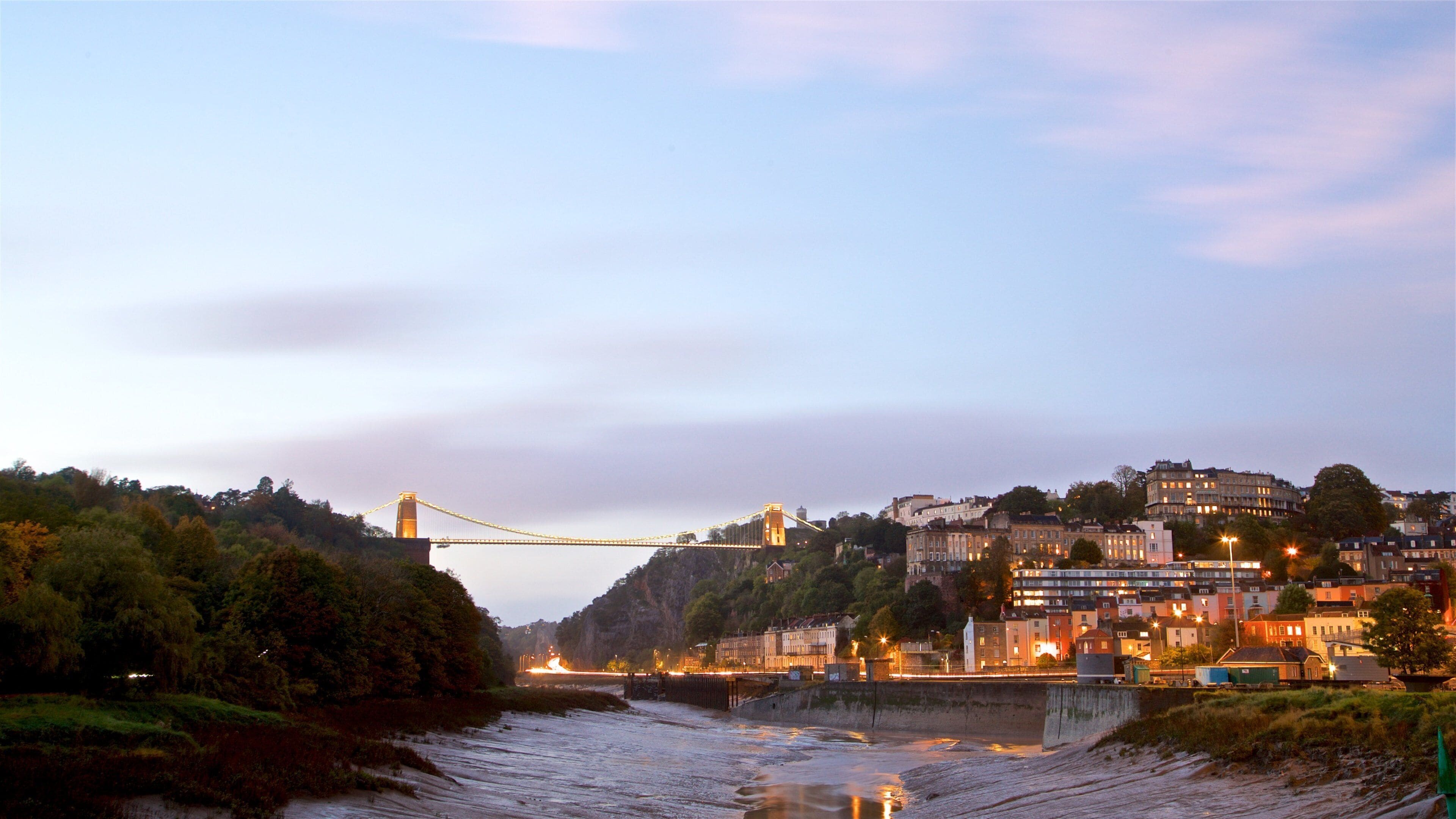 A suspension bridge over a river, with buildings and trees in the background.