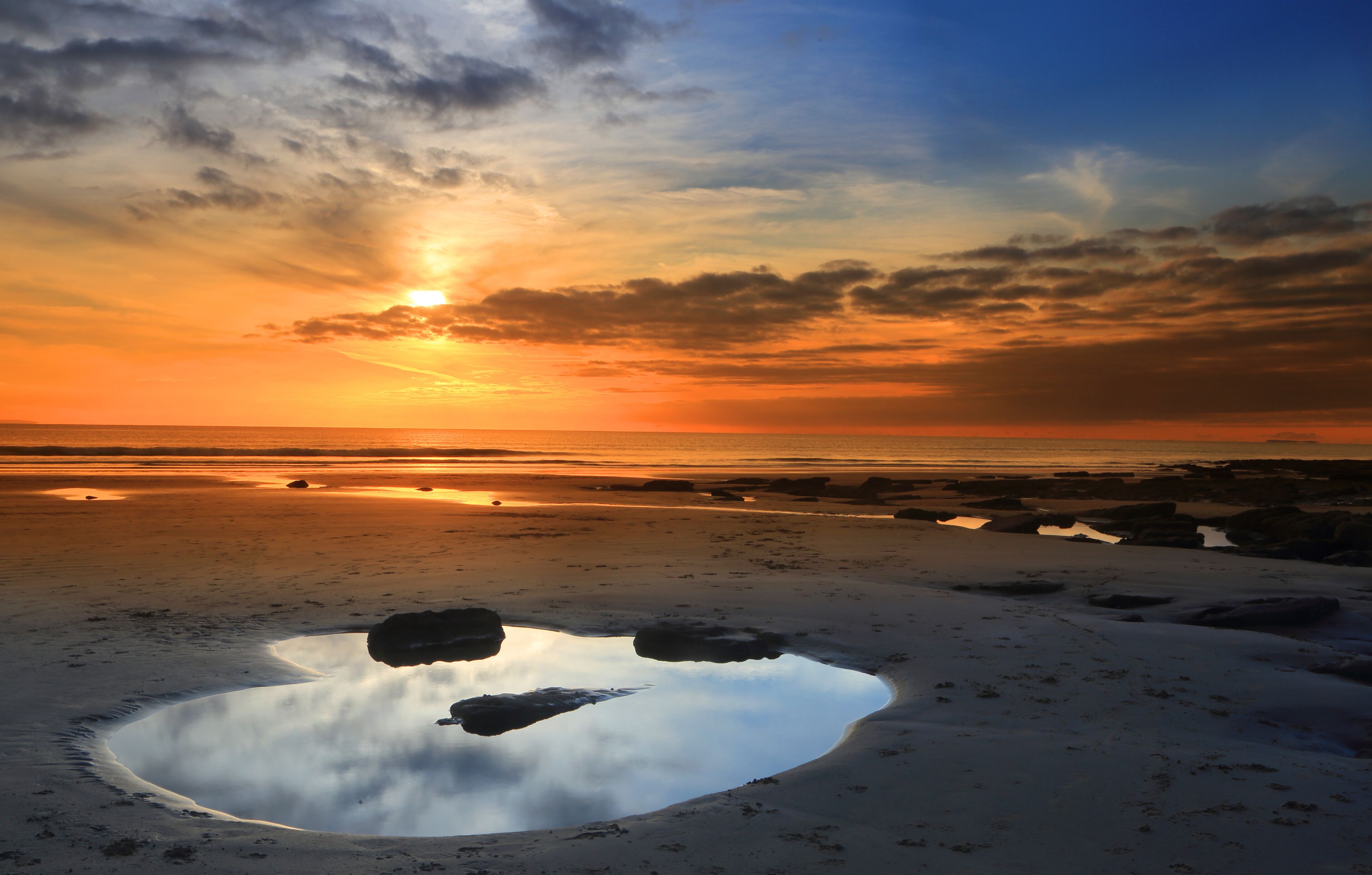 A sunset over a beach with a reflective puddle and scattered rocks.