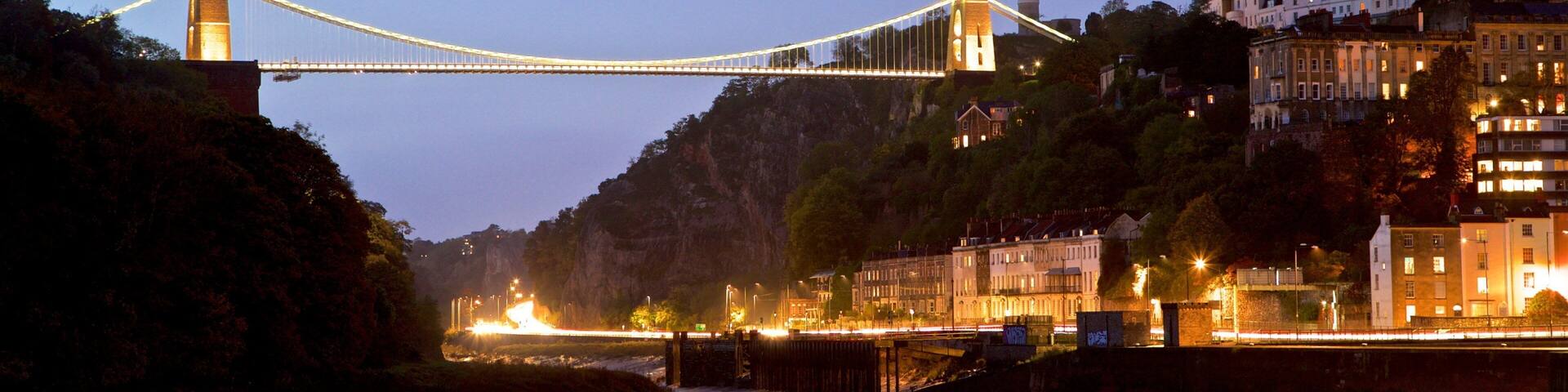 A suspension bridge over a river at dusk with city buildings and a hill in the background.