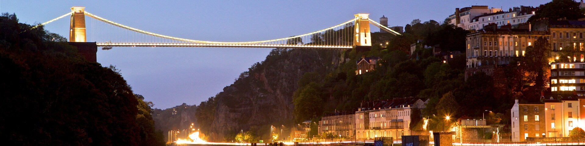 Un puente colgante sobre un río al atardecer, con edificios de la ciudad y una colina al fondo.