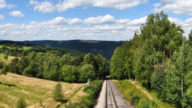 A railway track winding through a hilly landscape with dense forests and open fields under a partly cloudy sky.
