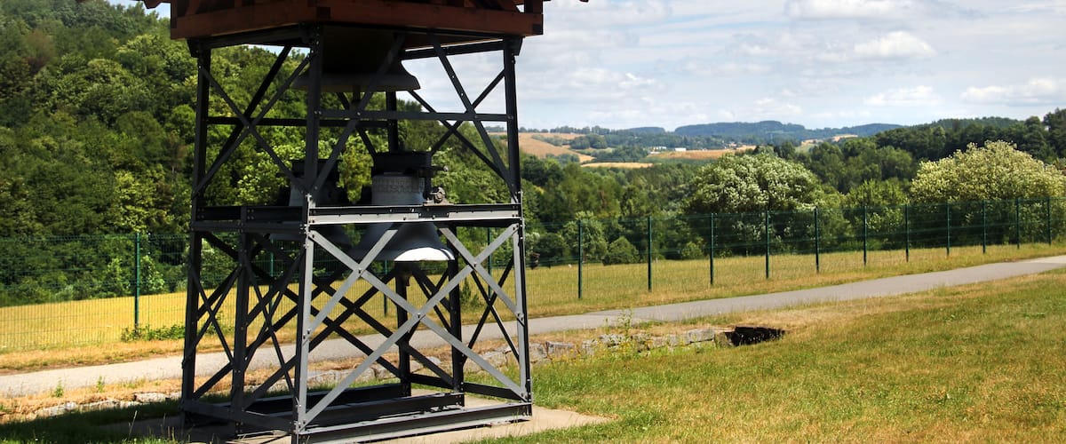 A wooden observation tower with a metal frame, surrounded by a fenced area with a pathway, set against a backdrop of green hills and a clear sky.