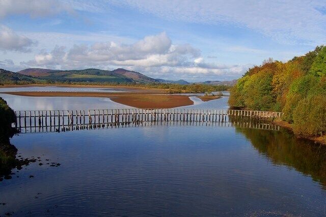 A calm river with a wooden bridge, surrounded by hills and autumn-colored trees.