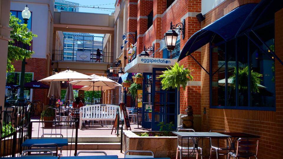 An outdoor cafe with tables and umbrellas, brick buildings, and hanging plants.