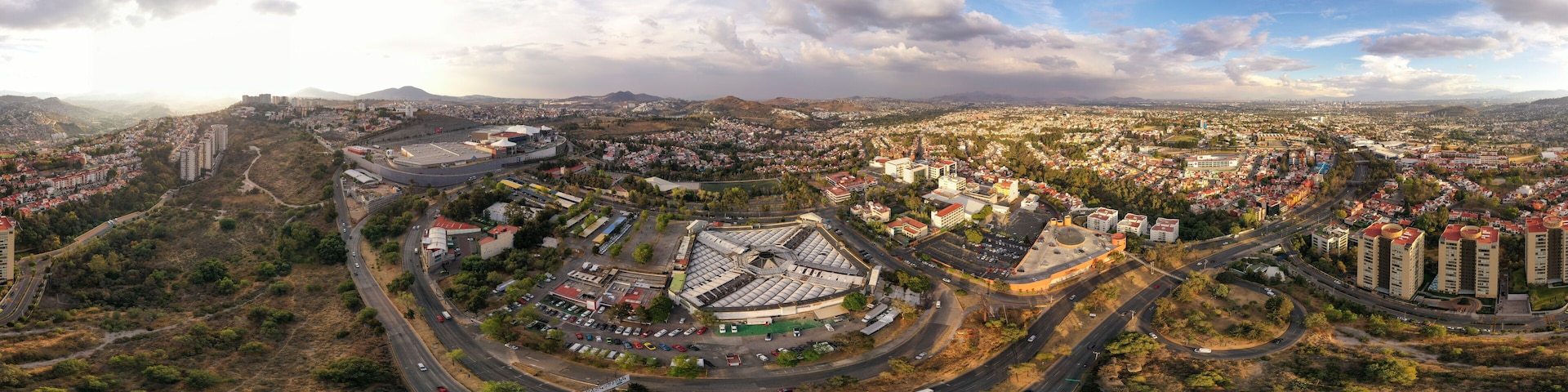A panoramic view of a city with a mix of residential and commercial buildings, a large roundabout, and surrounding hills.