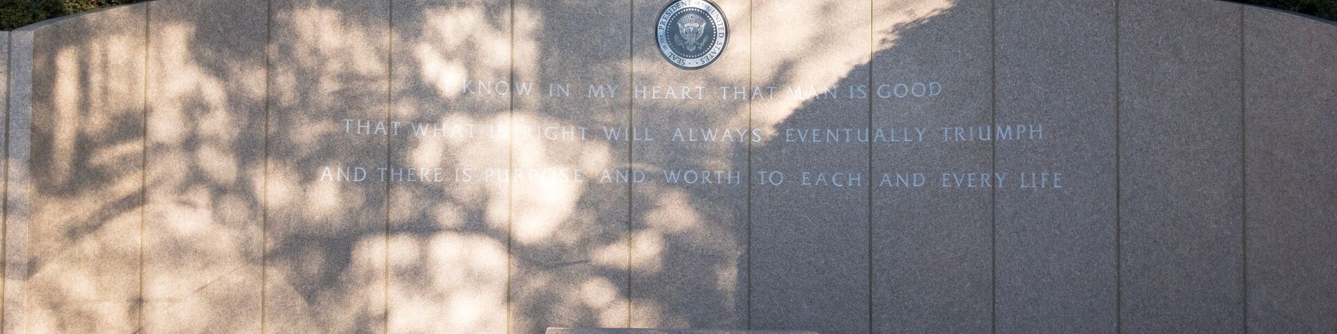 Un muro conmemorativo con inscripciones y un sello, rodeado de árboles.