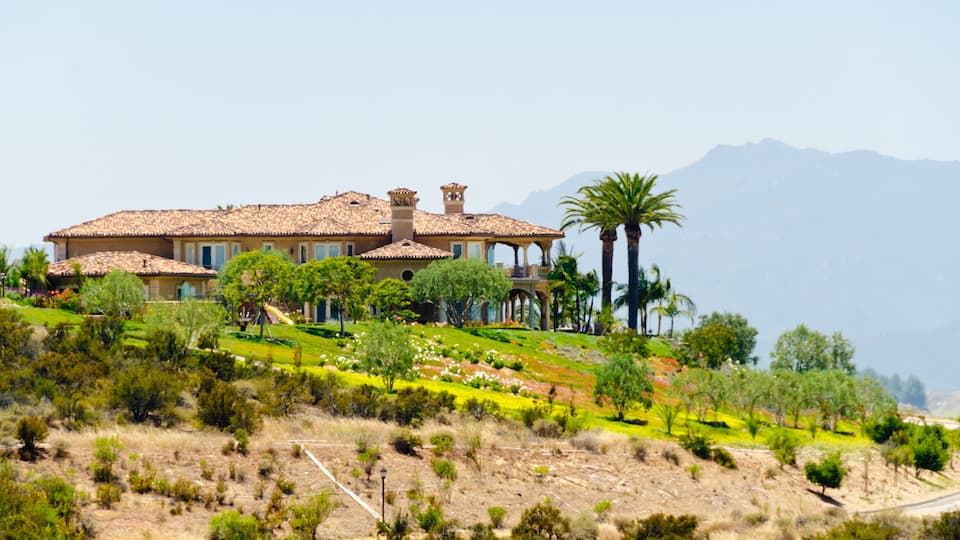 A large house with a tiled roof, surrounded by a landscaped garden with palm trees and a hilly landscape.