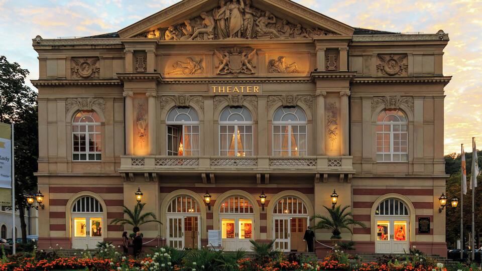 A grand theater with ornate sculptures and columns, surrounded by a well-maintained garden with vibrant flowers.