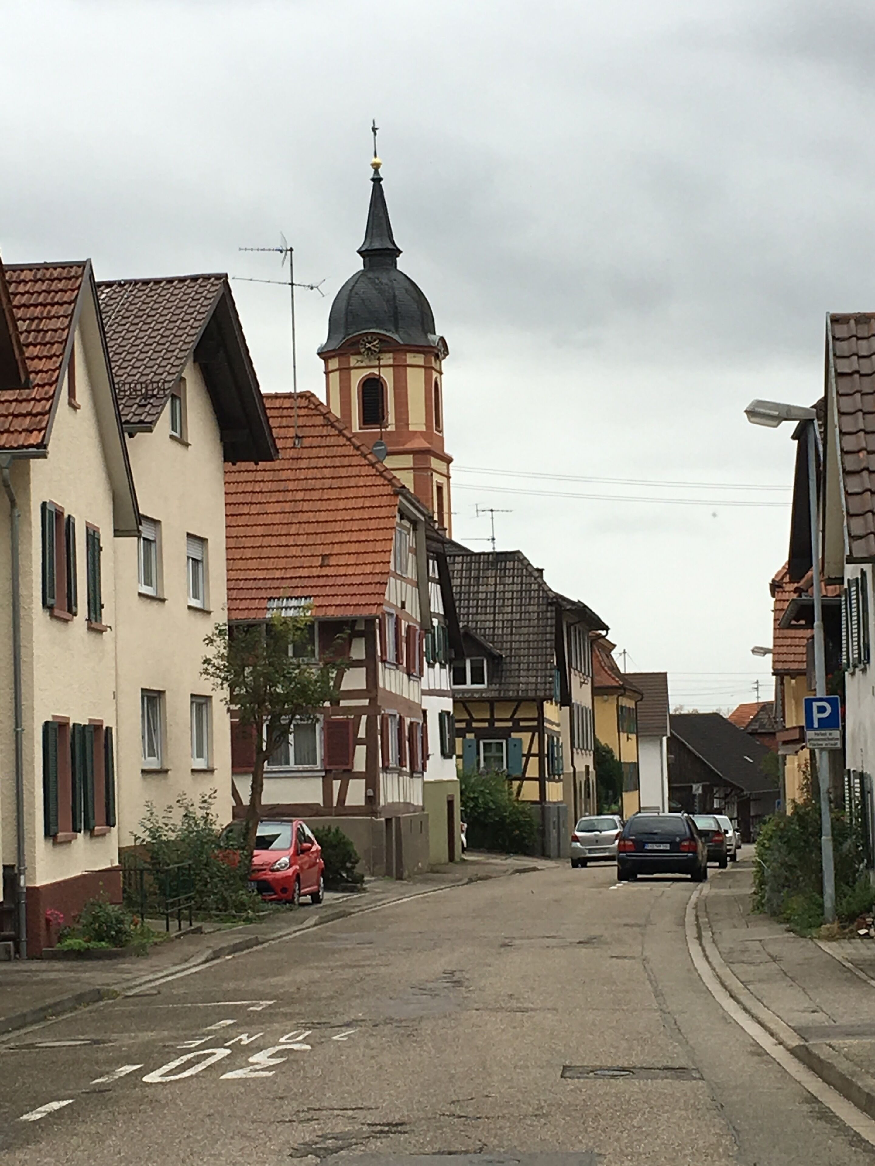 A narrow street lined with traditional half-timbered houses, a church with a clock tower, and parked cars.