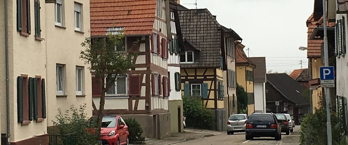 A narrow street lined with traditional half-timbered houses, a church with a clock tower, and parked cars.