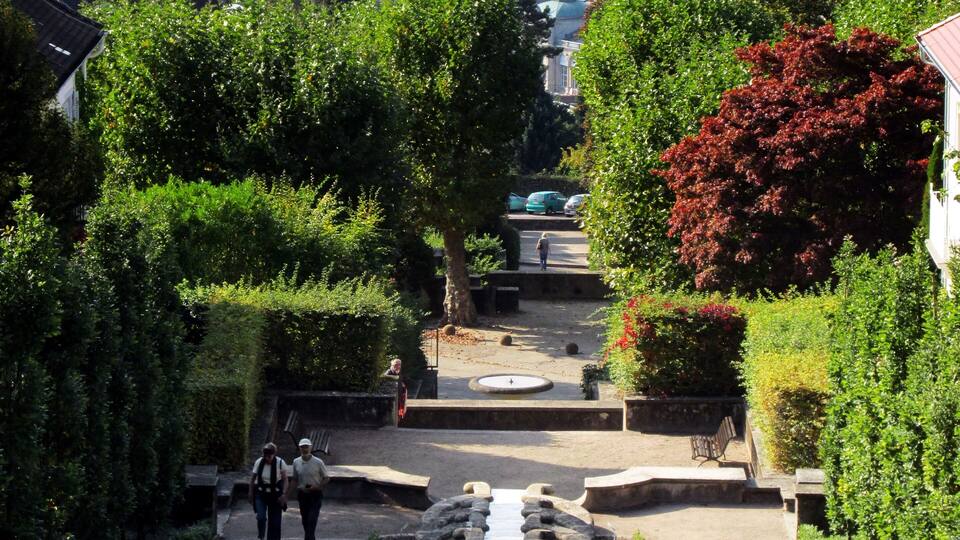 A historic town square with a central fountain, surrounded by lush greenery and traditional architecture.