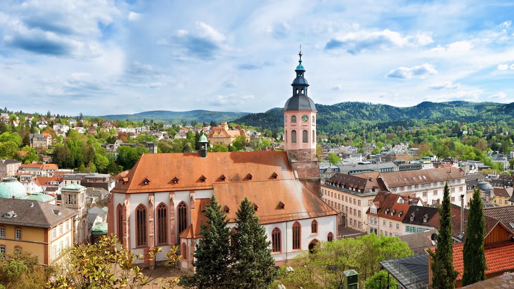Eine Stadt mit einem markanten Kirchengebäude, das einen hohen Turm und eine Uhr zeigt, umgeben von üppiger Vegetation und sanften Hügeln im Hintergrund.