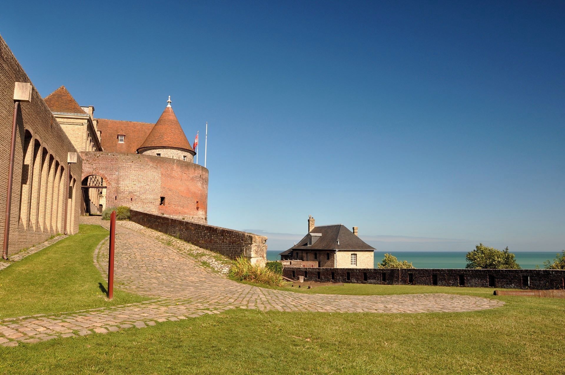 A historic castle with a round tower, a paved pathway, and a view of the sea.