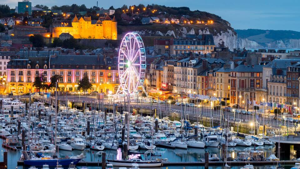 A harbor with numerous boats, a prominent Ferris wheel, and historic buildings with a castle on a hill in the background.
