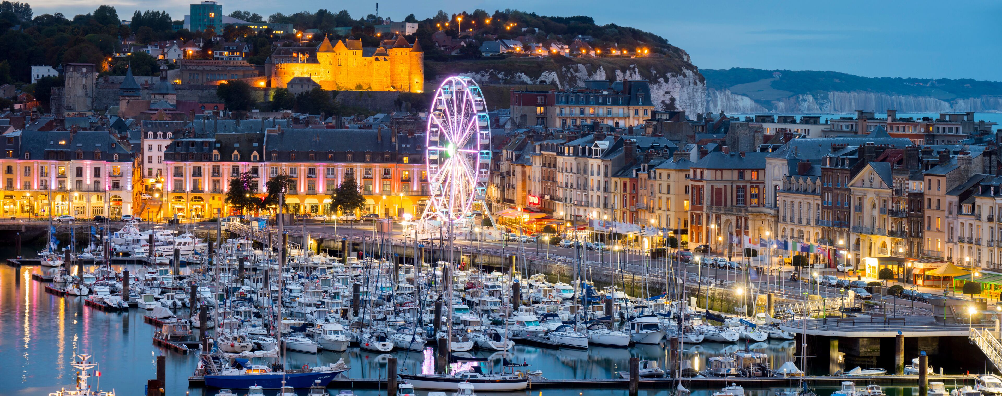 A harbor with numerous boats, a prominent Ferris wheel, and historic buildings with a castle on a hill in the background.