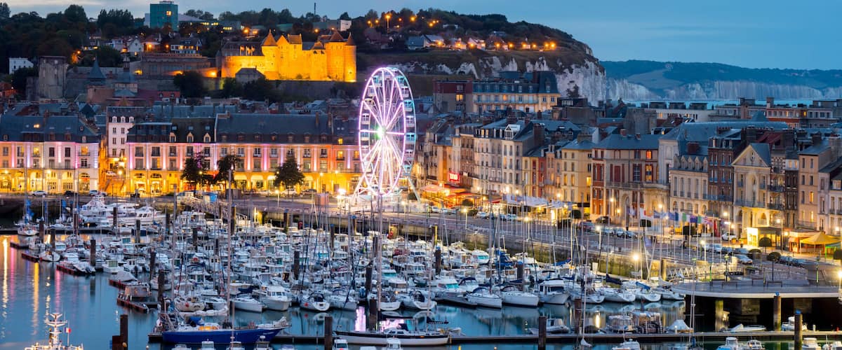 A harbor with numerous boats, a prominent Ferris wheel, and historic buildings with a castle on a hill in the background.