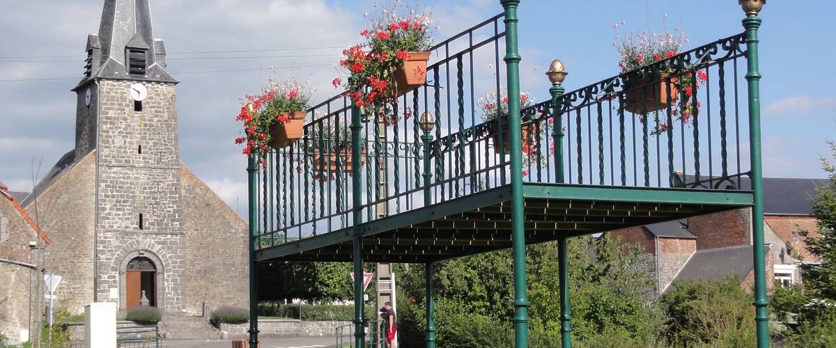 A green metal bridge with flower pots, a stone church with a clock tower, and a clear blue sky.