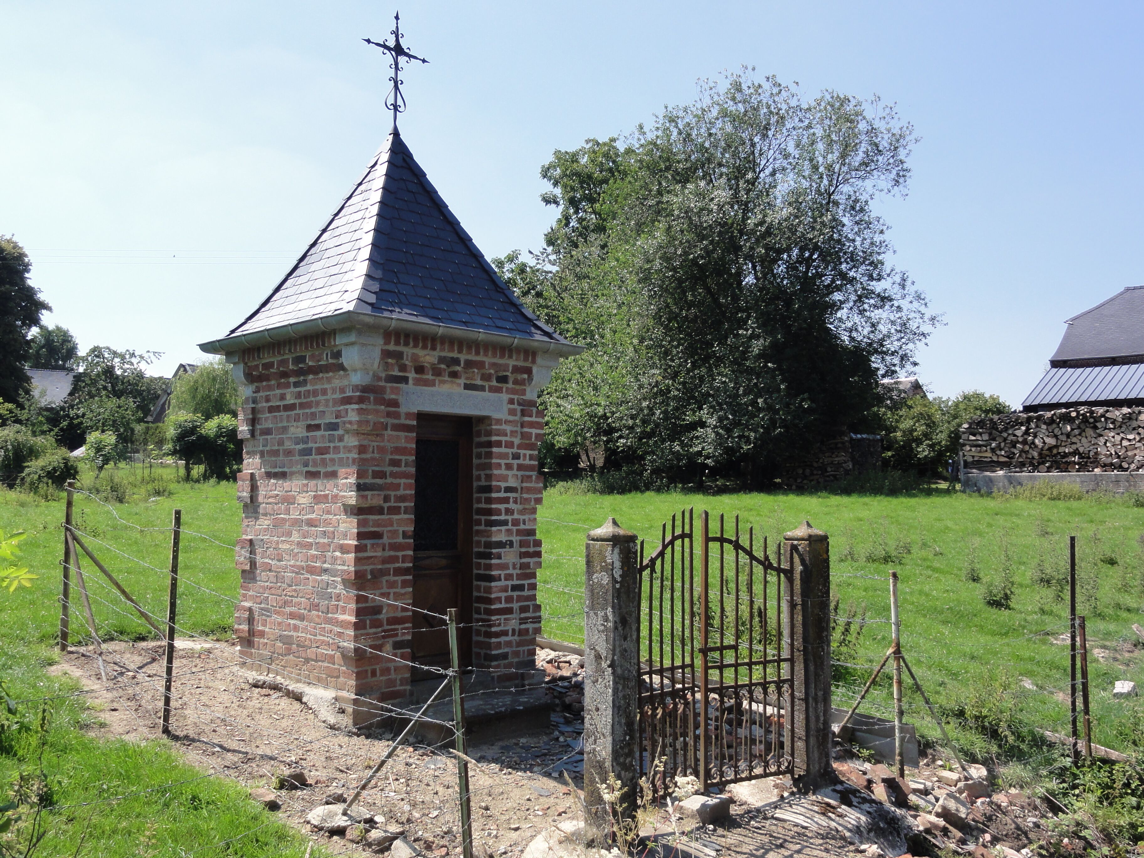 A small brick structure with a pointed roof and a weather vane, surrounded by a metal gate and a grassy field.