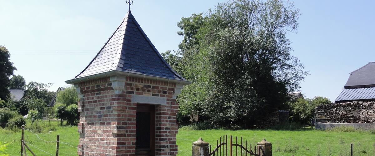 A small brick structure with a pointed roof and a weather vane, surrounded by a metal gate and a grassy field.